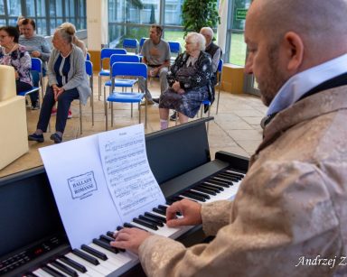 Ogród zimowy. Kierownik Arkadiusz Wanat gra na pianinie. Ma strój z epoki romantyzmu. W ogrodzie siedzą seniorzy.