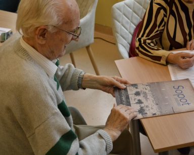 Sala. Przy stoliku siedzi senior. Przed nim kalendarz ze zdjęciem plaży i napisem Sopot 2022-2023.