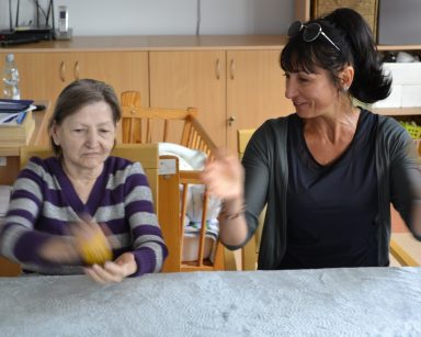 Sala. Przy stole siedzi terapeutka Ania Rzepczyńska i seniorka. Ćwiczą ręce.
