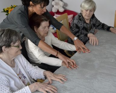 Sala. Przy stole siedzą seniorzy. Obok stoi terapeutka Ania Rzepczyńska. Ćwiczą ręce.