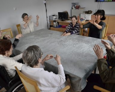 Sala. Przy stole siedzi terapeutka Ania Rzepczyńska i seniorzy. Ćwiczą ręce.