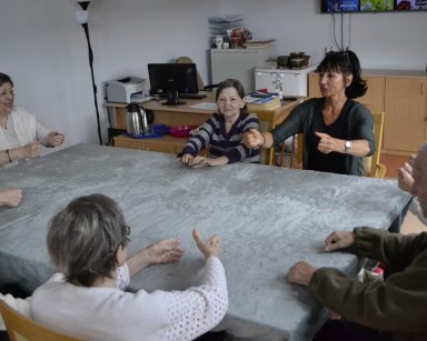 Sala. Przy stole siedzi terapeutka Ania Rzepczyńska i seniorzy. Ćwiczą ręce.