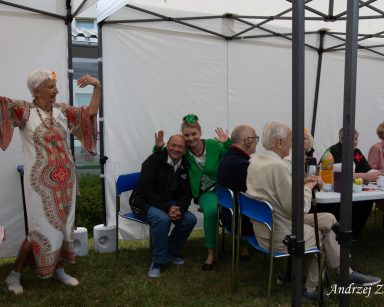 Festyn. Na świeżym powietrzu otwarte namioty. Seniorzy, goście, wolontariusze i pracownicy jedzą, rozmawiają, tańczą.