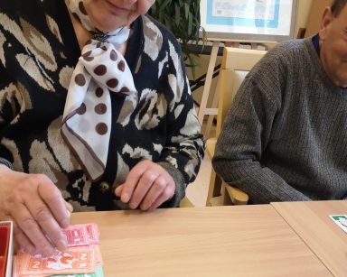 Sala. Przy stoliku siedzą seniorka i senior. Grają w monopoly.