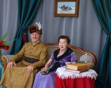 Salonik. Senior i seniorka pozują do zdjęcia w strojach z epoki romantyzmu. Siedzą na sofie.