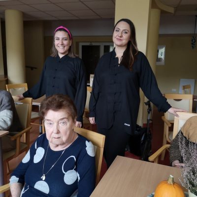 Sala. Przy stolikach siedzi czworo seniorów. Za nimi stoją fizjoterapeutki Martyna Józefczyk i Paulina Winczura.