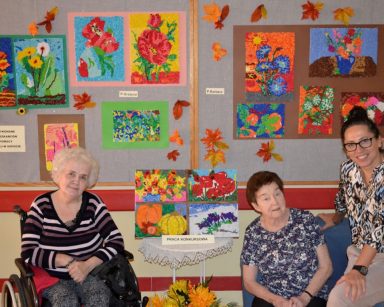 Dwie seniorki i terapeutka Magdalena Poraj-Górska pozują do zdjęcia. Za nimi na tablicy wiszą kolorowe obrazy.