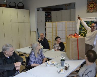 Świetlica. Seniorzy siedzą przy stołach. Na blacie ogromny prezent. Terapeutka Magdalena Poraj-Górska trzyma nożyczki.