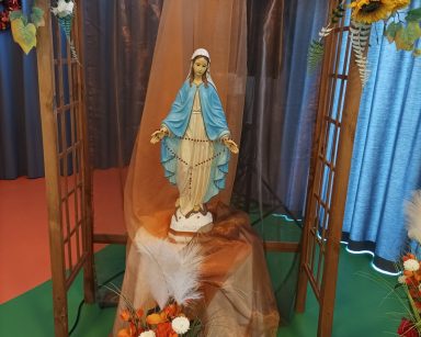 Sala. Drewniana pergola ozdobiona kwiatami. W środku figurka Matki Boskiej z różańcem.