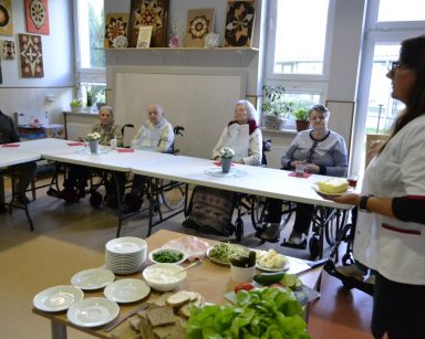 Sala. Seniorzy siedzą przy stołach. Na środku stoi dietetyczka Karolina Gorzkiewicz. Prowadzi zajęcia.