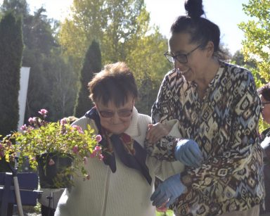 Pogodny dzień. Ogród. Na pierwszym planie terapeutka Magdalena Poraj-Górska i seniorka. Niosą kwiaty.