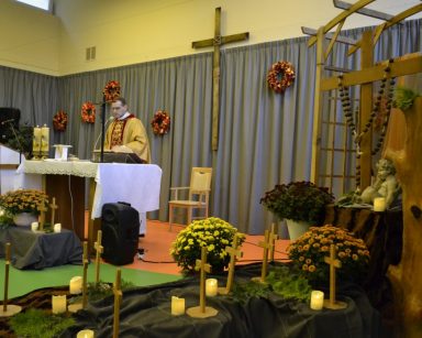 Sala. Ksiądz Tomasz Kosewski stoi przy ołtarzu. Odprawia mszę. Dekoracja z chryzantem, drewnianych krzyży i świeczek.
