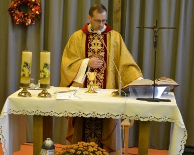 Ksiądz Tomasz Kosewski stoi przy ołtarzu. Przed ołtarzem dekoracja. Chryzantemy, świeczki, drewniane krzyże.