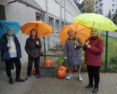 Pochmurny dzień. Przed budynkiem stoją cztery seniorki. Mają kolorowe parasole.