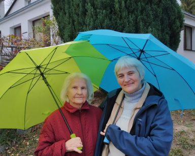 Pochmurny dzień. Na świeżym powietrzu dwie seniorki. Uśmiechają się. Mają kolorowe parasole.
