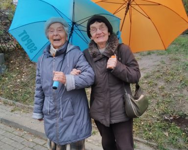 Pochmurny dzień. Na świeżym powietrzu dwie seniorki. Śmieją się. Mają kolorowe parasole.