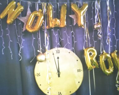 Dekoracja. Duży okrągły zegar ze wskazówkami. Wskazuje minutę przed 12. Wiszą bombki, lampki, napis z balonów Nowy rok.