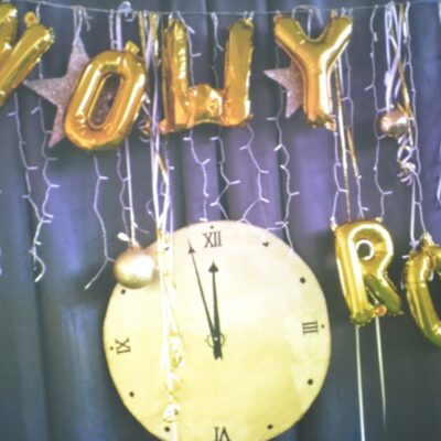 Dekoracja. Duży okrągły zegar ze wskazówkami. Wskazuje minutę przed 12. Wiszą bombki, lampki, napis z balonów Nowy rok.
