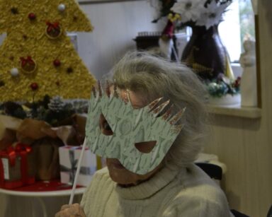 Świetlica. Przy stole siedzi seniorka. Przykłada do twarzy karnawałową maskę.