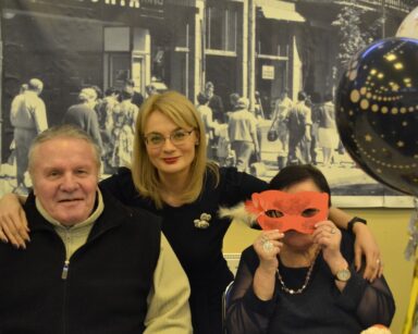 Sala. Dyrektorka Agnieszka Cysewska pozuje do zdjęcia z seniorem i seniorką. Seniorka trzyma karnawałową maskę.
