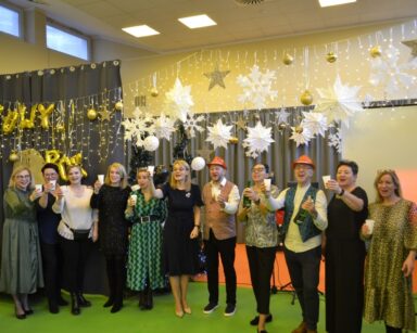 Sala. Sylwestrowe dekoracje. Dyrektorka Agnieszka Cysewska i pracownicy stoją w rzędzie. Wznoszą toast szampanem.