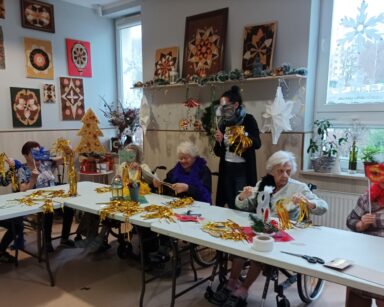 Świetlica. Seniorki, terapeutka Magdalena Poraj-Górska i wolontariuszka pokazują karnawałowe maski i złote kity.