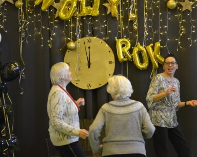 Sala. Sylwestrowa dekoracja. Duży zegar, balony, napis Nowy rok. Trzy kobiety tańczą.