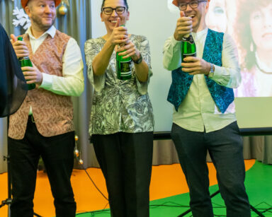 Sala. Karnawałowe dekoracje. Troje pracowników trzyma butelki z szampanem. Śmieją się.