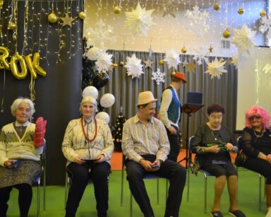 Sala. W rzędzie na krzesłach siedzą senior i cztery seniorki. Mają różne rekwizyty: rękawice, korale, perukę, kapelusz, okulary.