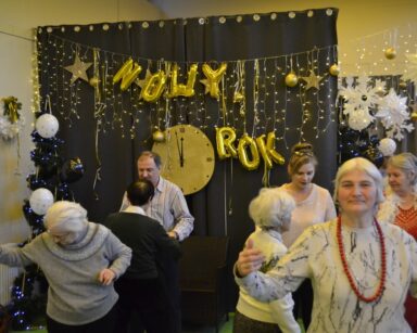 Sala. Sylwestrowa dekoracja. Duży zegar, balony, napis Nowy rok. Seniorzy i pracownicy tańczą.