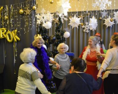 Sala. Sylwestrowa dekoracja. Duży zegar, balony, napis Nowy rok. Seniorzy, pracownicy, wolontariusze tańczą.