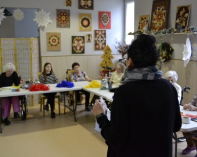 Świetlica. Terapeutka Magdalena Poraj-Górska prowadzi spotkanie. Przy stołach siedzą seniorki i wolontariuszka.