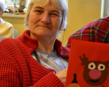 Sala. Seniorka pokazuje świąteczną kartkę z reniferem. Uśmiecha się.