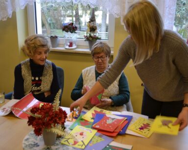 Sala. Na pierwszym planie zastępczyni dyrektora Ilona Gajewska i dwie seniorki oglądają świąteczne kartki.