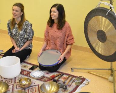 Sala. Zajęcia dźwiękoterapii. Na podłodze siedzą dwie kobiety. Przed nimi misy nepalskie, obok gong.