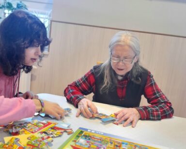 Świetlica. Seniorka i wolontariuszka siedzą przy stole. Układają puzzle.
