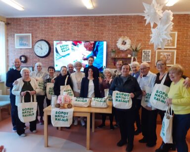 Sala. Seniorzy, pracownicy i goście pozują do grupowego zdjęcia. Trzymają torby z napisem Sopot naturalnie. Śmieją się.