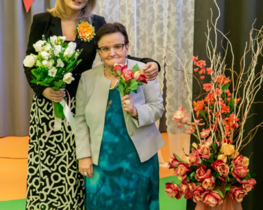 Sala. dyrektorka Agnieszka Cysewska i seniorka pozują do wspólnego zdjęcia. Uśmiechają się. Każda trzyma bukiet kwiatów.