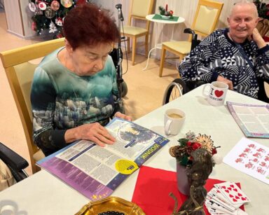 Świetlica dwoje seniorów siedzi przy stole. Seniorka czyta gazetę z horoskopem. Na stole karty, gazety, kawa, ciastka.