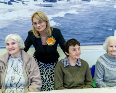 Sala. Przy stole siedzą trzy seniorki. Za nimi stoi dyrektorka Agnieszka Cysewska. Uśmiechają się, pozują do zdjęcia.