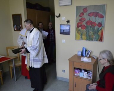 Pokój. Wizyta duszpasterska księdza Tomasza Kosewskiego. Seniorka siedzi na łóżku.