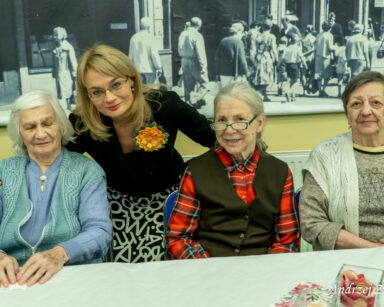 Sala. Przy stole siedzą trzy seniorki. Za nimi stoi dyrektorka Agnieszka Cysewska. Uśmiechają się, pozują do zdjęcia.