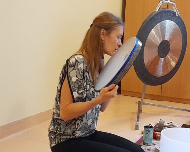 Sala. Kobieta prowadzi zajęcia dźwiękoterapii. Obok zawieszony gong.