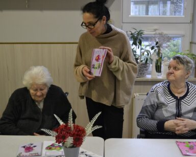 Świetlica. Przy stole siedzą dwie seniorki. Obok stoi terapeutka Magdalena Poraj-Górska. Pokazuje laurkę.