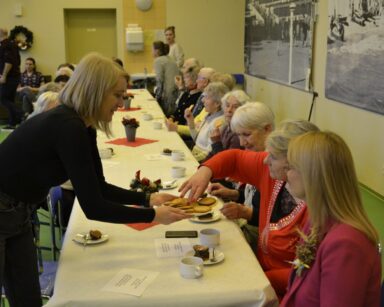 Sala. Seniorzy i pracownicy przy stole. Na pierwszym planie zastępczyni Ilona Gajewska częstuje seniorkę ciastkiem.