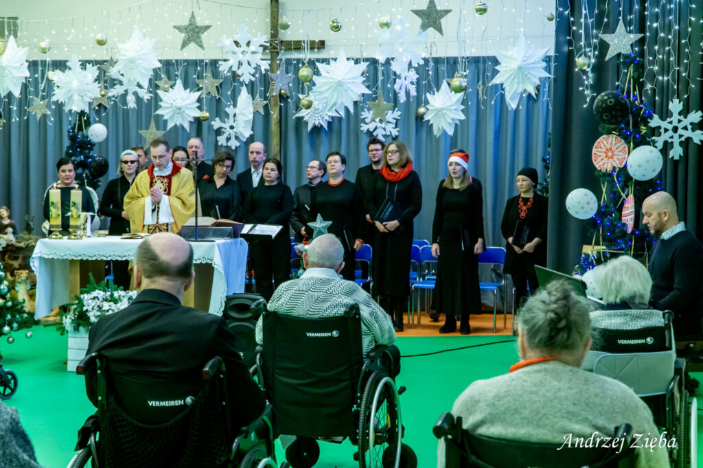 Sala. Ksiądz Tomasz Kosewski odprawia mszę. Przed nim siedzą seniorzy. Za nim stoją chórzyści.