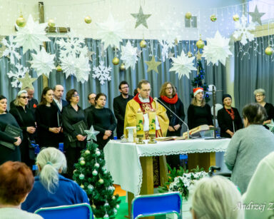 Sala. Zimowe dekoracje. Ksiądz Tomasz Kosewski odprawia mszę. Przed nim siedzą seniorzy. Za nim stoją chórzyści.