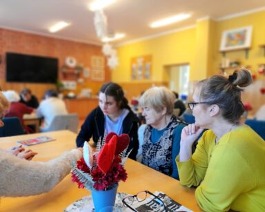 Sala. Na pierwszym planie dwie seniorki, terapeutka Beata Brzozowska, młoda wolontariuszka. Siedzą razem przy stole.
