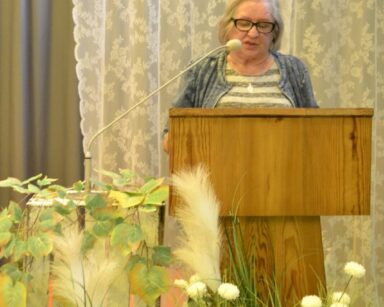 Sala. Seniorka stoi na mównicy. Czyta. Przed mównicą dekoracja z białych kwiatów i zielonych roślin.