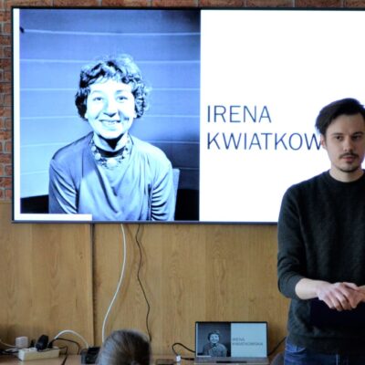 Sala. Szymon Ropel stoi przy telewizorze. Na ekranie czarno-białe zdjęcie aktorki Ireny Kwiatkowskiej.